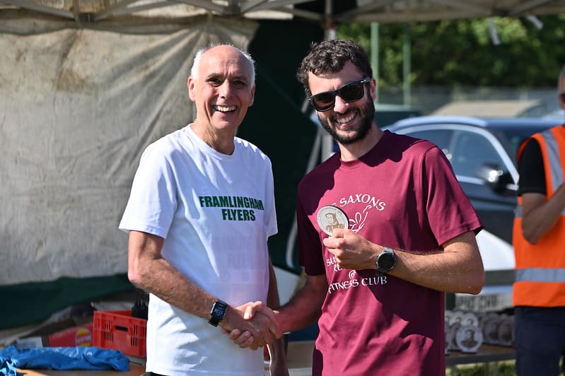 Two men wearing running t-shirts shaking hands one holding a medal and smiling at the camera