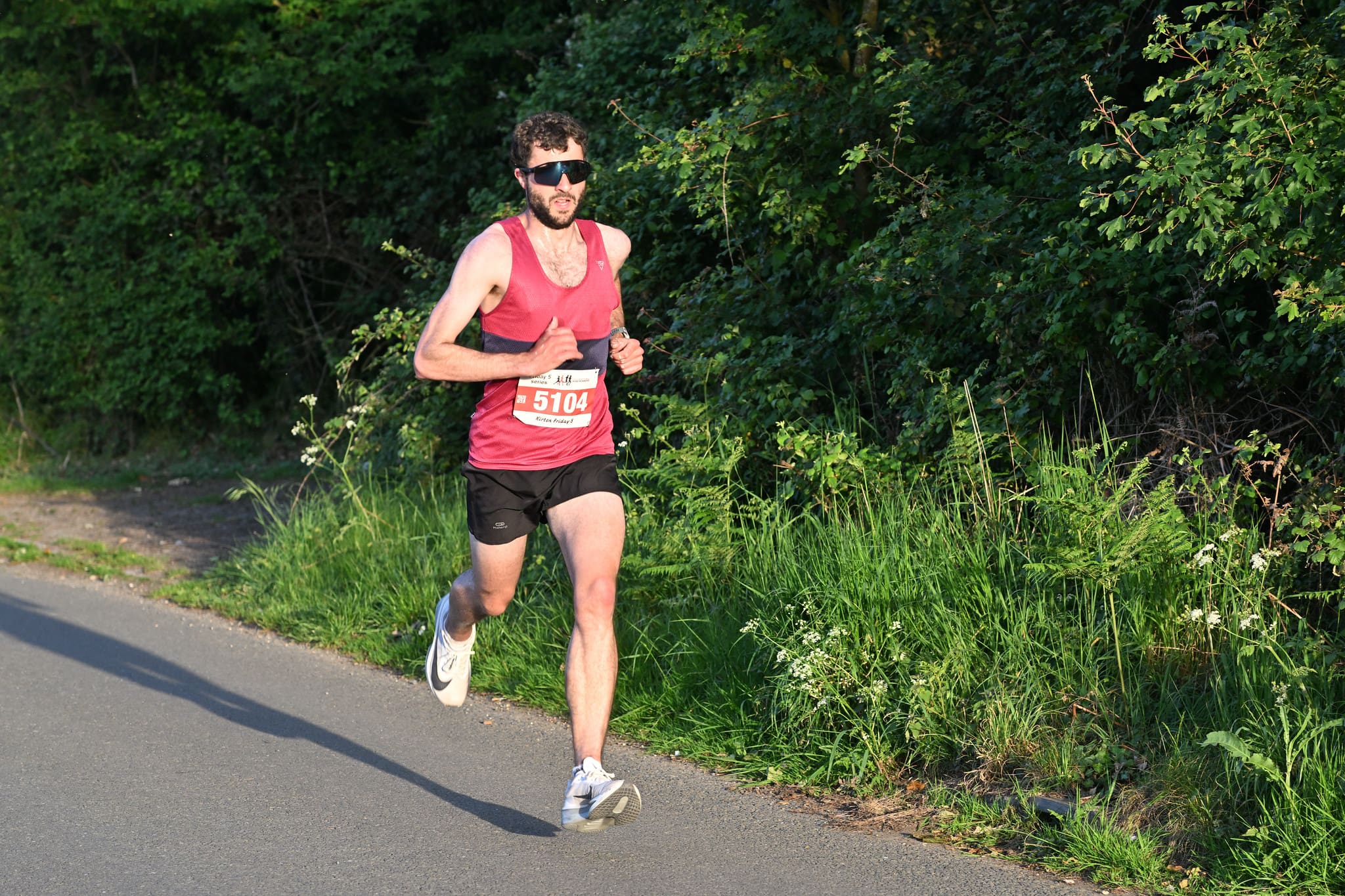 Man wearing a burgundy running vest and black shorts running down a road in the sunshine